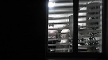 Peeping. Voyeur. Neighbor pervert Hidden cam in evening on street looks out window as nude super-sexy neighbor in kitchen prepares dinner for her husband. nude in public. nude at home. Family.  Outdoor